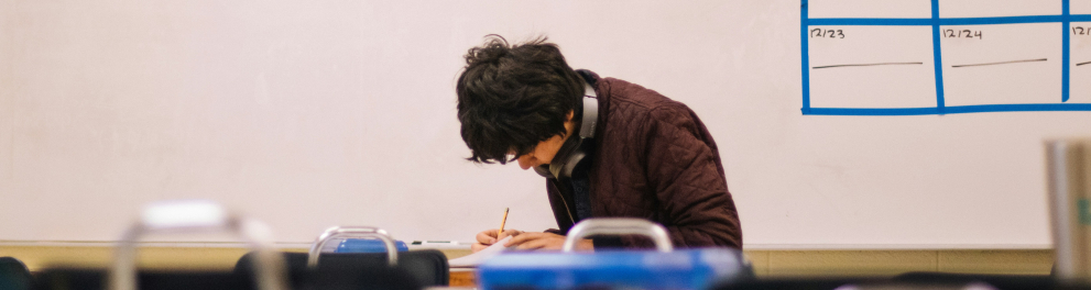 Um estudante sentado em uma carteira em uma sala de aula de cabeça baixa olhando para um caderno