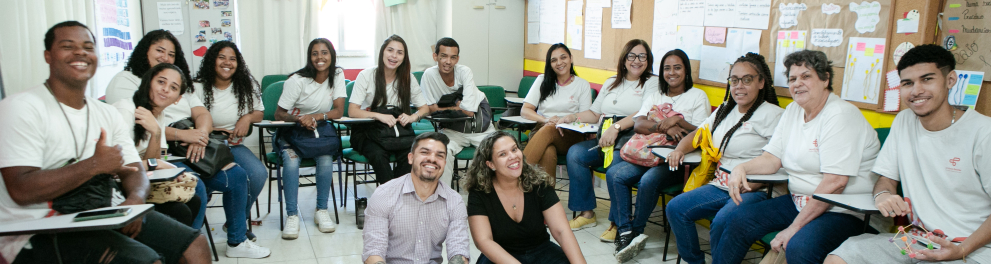 Grupo de estudantes em semi-círculo, sentados em uma sala de aula. No chão, ao centro, há duas pessoas sentadas, um homem e uma mulher.