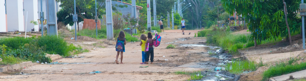 Ao longe, as crianças vão a pé para a escola em uma rua não asfaltada e com esgotão a céu aberto