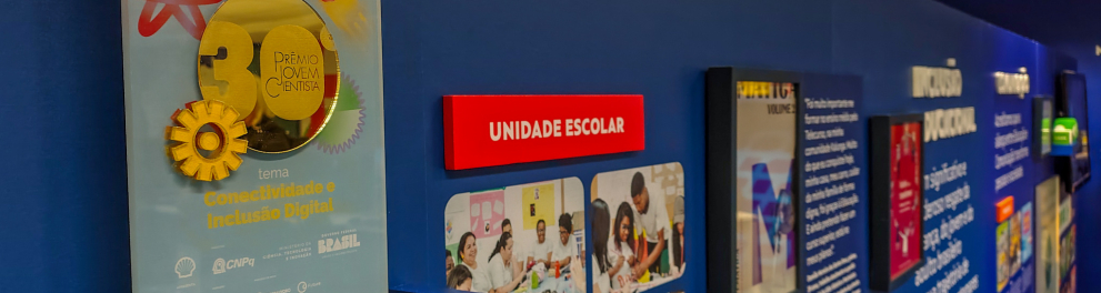 Vista de um painel expositivo com o troféu do Prêmio Jovem Cientista com o tema “Conectividade e Inclusão Digital”. A parede apresenta iniciativas educacionais da Fundação Roberto Marinho, sob o eixo “Unidade Escolar”, com fotos de estudantes e professores em atividades escolares e de tecnologia. Ao fundo, são visíveis outros segmentos temáticos como "Inclusão Vocacional" e "Trabalho".