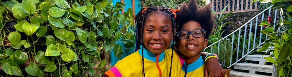 Duas crianças estão abraçadas e sorrindo em um jardim cheio de plantas verdes. Estão vestindo os mesmos macacões amarelos com detalhes coloridos. Atrás delas há uma escada de azulejos e muitas plantas ao redor. Em frente, sobre uma bancada de madeira, há vasos e mudas com etiquetas, indicando um espaço de cultivo e aprendizado sobre plantas.