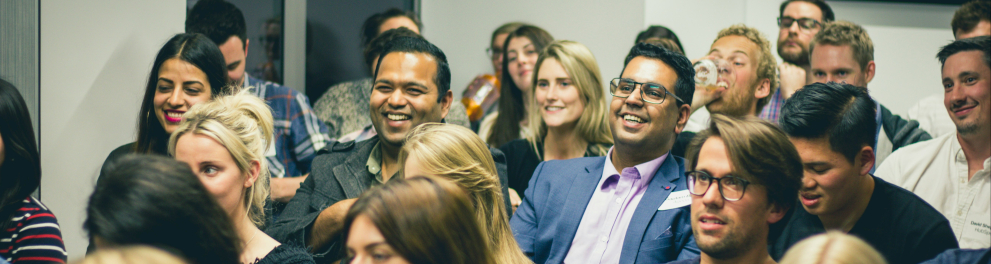 Fotografia colorida de um grupo de pessoas sentadas em um auditório ou sala de reuniões, participando de um evento. A maioria das pessoas está sorrindo ou rindo, sugerindo que estão assistindo a algo divertido ou interessante. Há uma diversidade de rostos, com diferentes tons de pele, gêneros e estilos. Todos estão vestidos de maneira casual ou social leve. O ambiente é fechado, com paredes brancas e iluminação artificial. Ao fundo, há uma televisão ou tela preta montada na parede.