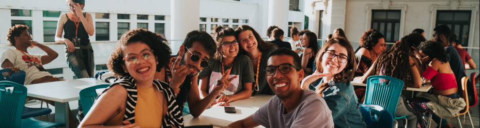 A imagem mostra um grupo de jovens sentados em mesas e alguns deles posam para a foto