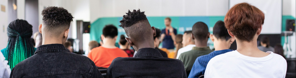 A imagem mostra um grupo de jovens sentados em cadeiras enfileiradas, assistindo a uma apresentação em um ambiente interno, semelhante a uma sala de aula ou auditório. A foto é tirada de trás, mostrando principalmente as costas das pessoas. Três jovens na fileira da frente se destacam: dois com jaquetas pretas e cortes de cabelo estilizados, e um com camiseta branca e cabelo castanho-avermelhado. Ao fundo, há um homem em pé segurando um microfone, aparentemente falando ao público. Atrás dele, há uma tela de