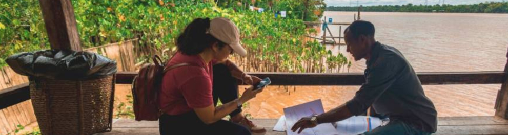 Dois adultos sentados sob um quiosque de madeira com cobertura de palha, à beira de um rio em área de floresta amazônica. À esquerda, uma mulher com boné claro, mochila vermelha e camiseta rosa segura um celular e observa um papel. À direita, um homem de bermuda jeans e camisa escura aponta para mapas ou documentos sobre o banco. O ambiente é ribeirinho, com vegetação densa e o rio ao fundo, transmitindo um clima de conversa e troca de saberes em meio à natureza.