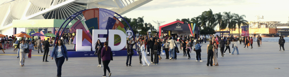 A imagem mostra uma praça com diversas pessoas transitando e um edíficio branco ao fundo, que é o Museu do Amanhã.