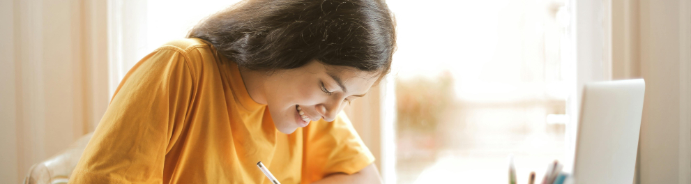 A imagem mostra uma jovem sentada escrevendo algo em um caderno e sorrindo