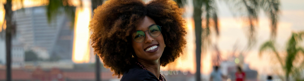 Uma mulher negra sorrindo e sentada e posando de lado para uma foto em frente ao espelho d'água do Museu do Amanhã