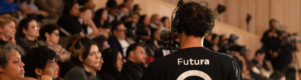 Plateia de um auditório lotado, com pessoas sentadas e atentas à programação. Em primeiro plano, de costas, está um cinegrafista usando fones de ouvido e camiseta preta com o logotipo do Canal Futura, que grava o evento. Algumas pessoas do público seguram celulares e objetos coloridos nas mãos. O ambiente tem iluminação suave e paredes revestidas de madeira clara.