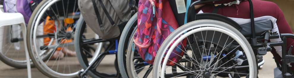 Uma foto que foca nas rodas e nos encostos de três cadeiras de rodas em um espaço externo. Duas pessoas estão sentadas nas cadeiras, mas apenas a parte de baixo de seus corpos aparece. Uma mochila colorida, com bolinhas, está pendurada no encosto da cadeira de rodas em primeiro plano.