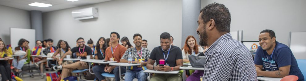 Na imagem, um grupo de jovens universitários está reunido em uma sala de aula moderna. Eles estão sentados em cadeiras com rodinhas e pequenas mesas acopladas, formando um semicírculo. Todos prestam atenção e sorriem para um homem que está à frente, sentado em uma cadeira vermelha, conversando de forma descontraída. A sala é clara, com paredes brancas, ar-condicionado e iluminação de teto. O clima é leve e de troca, com os estudantes demonstrando interesse e engajamento na atividade.