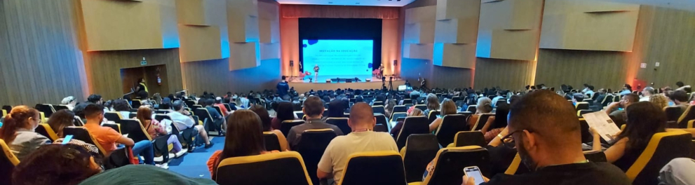 A imagem mostra um auditório com muitas pessoas e, ao fundo, pessoas apresentam algo em um telão