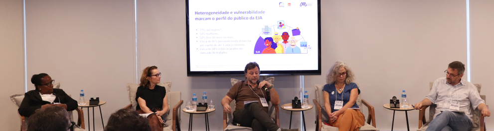 Um grupo de cinco pessoas está sentado em cadeiras dispostas em semicírculo, participando de um painel de discussão em um auditório iluminado. No centro, atrás dos debatedores, há uma tela grande com um slide projetado que traz o título: “Heterogeneidade e vulnerabilidade marcam o perfil do público da EJA”. O slide apresenta alguns dados estatísticos e uma ilustração colorida de pessoas diversas. À frente da tela, os participantes estão sentados com microfones e garrafas de água sobre pequenas mesas de apoi