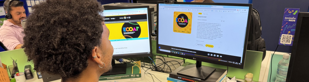 Na imagem, um jovem está sentado em frente a dois monitores de computador em um ambiente de escritório. Ele navega em uma página sobre o Edital ECOAR, visível na tela da direita, enquanto o monitor da esquerda exibe a identidade visual colorida do projeto. Na mesa, há livros empilhados que servem de suporte para os monitores, teclado, celular e materiais de papelaria. Ao fundo, outros colegas de trabalho também utilizam seus computadores, criando um clima colaborativo de escritório.