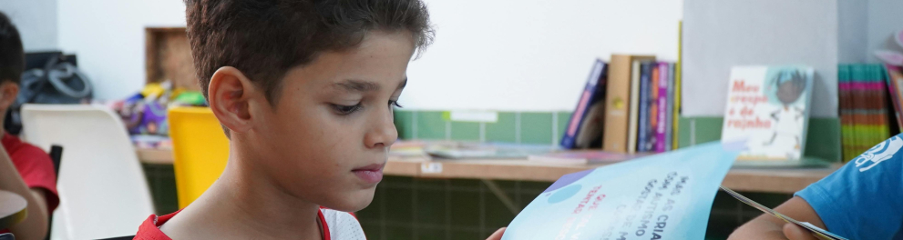 A imagem mostra uma criança em uma escola, sentada em uma carteira escolar e segurando e lendo um livro didático com as mãos. 
