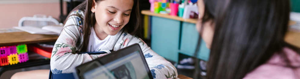A imagem mostra crianças em uma sala de aula. Uma delas, uma menina, está sentada em uma mesa com um notebook em sua frente.