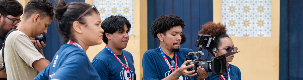 Um grupo de jovens está reunido em uma rua de paralelepípedos, em frente a uma parede amarela decorada com azulejos brancos e azuis. Todos vestem camisetas azul-marinho com crachás vermelhos pendurados no pescoço. No centro, um rapaz segura uma câmera de cinema, focado na filmagem, enquanto os colegas observam atentamente. A atmosfera é de aprendizado e trabalho em equipe durante uma atividade prática de gravação.