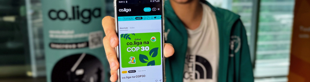 Na imagem, um jovem sorri enquanto exibe, em primeiro plano, a tela de seu celular que mostra o edital “co.liga na COP 30” com fundo verde e ilustrações coloridas. Ele usa uma jaqueta verde sobre uma camiseta branca e está em um ambiente interno iluminado, com banners da plataforma co.liga desfocados ao fundo. A foto transmite entusiasmo e engajamento juvenil com a iniciativa voltada à Conferência do Clima.