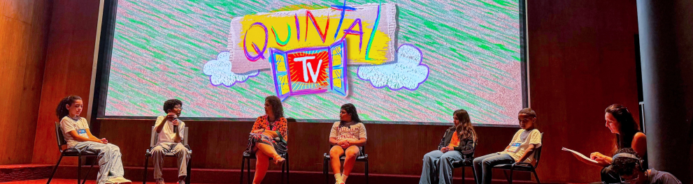 Seis pessoas estão sentadas em cadeiras no palco de um auditório, diante de uma grande tela que exibe o logotipo colorido da série Quintal TV, desenhado em estilo infantil, com fundo verde e nuvens brancas. Cinco são crianças e uma é adulta. Uma das crianças segura um microfone, enquanto uma mulher, à direita, lê um papel. Um cinegrafista, agachado, registra o momento com uma câmera. O ambiente é fechado, com paredes de madeira e carpete vermelho.