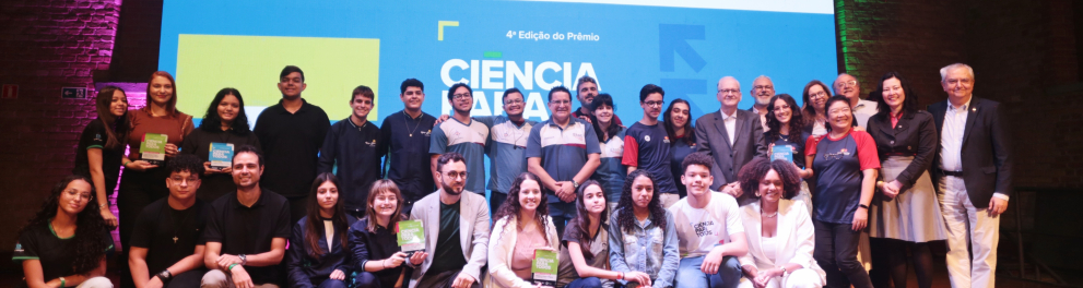 A imagem mostra um grupo maior, composto por estudantes, professores, organizadores e autoridades, reunidos sobre um palco de madeira. Ao fundo, está o telão com a identidade visual da 4ª edição do Prêmio Ciência Para Todos. O grupo está distribuído em dois níveis: parte em pé e parte sentada ou agachada. Algumas pessoas seguram placas ou troféus. As expressões são positivas, com muitos sorrindo. O ambiente conserva paredes de tijolos aparentes e iluminação colorida em tons de roxo e verde.