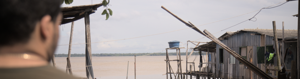 À beira de um rio de águas amarronzadas, casas de madeira sobre palafitas se estendem sobre o espelho d’água. Um homem observa a paisagem, parcialmente de costas. Pequenos barcos coloridos estão atracados e o céu nublado reforça o clima amazônico e ribeirinho.