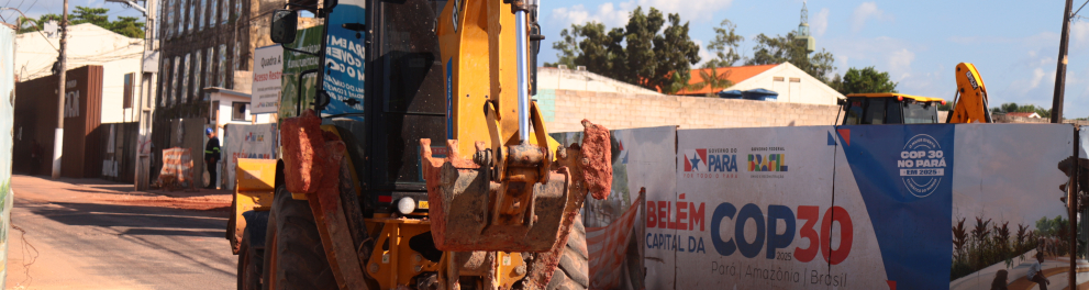 Uma retroescavadeira amarela realiza obras em uma rua de Belém, cercada por tapumes que exibem o logotipo do Governo do Pará e a frase “Belém, capital da COP 30 – Pará, Amazônia, Brasil”. O céu está azul e, ao fundo, há construções antigas e fios de energia cruzando a cena, indicando preparativos na cidade para receber o evento climático.