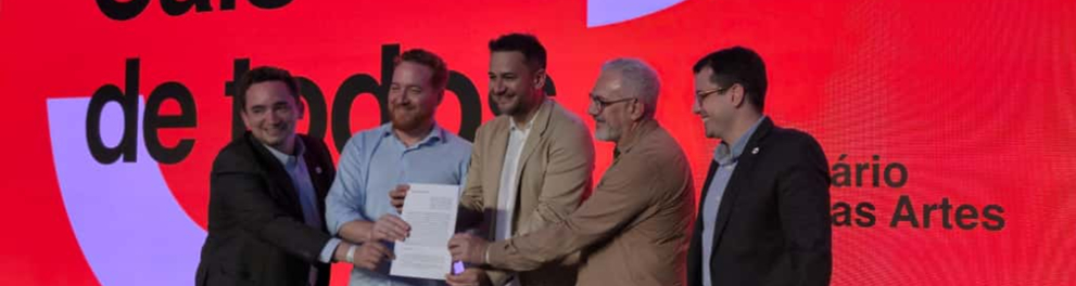 Cinco homens estão em pé sobre um palco, sorrindo e segurando juntos um documento, em gesto simbólico de assinatura de parceria. Eles estão diante de um grande painel digital vermelho com letras pretas e brancas que formam as palavras “Cais de todos” e parte do texto “Cais das Artes”. À frente deles há uma mesa com papéis. O ambiente tem iluminação de evento e atmosfera formal, sugerindo uma cerimônia de anúncio ou assinatura institucional.