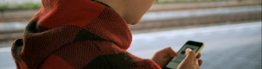 A foto mostra uma pessoa jovem de perfil, com cabelo loiro preso em um coque alto, usando um casaco xadrez em tons de vermelho e preto. Ela está em uma plataforma de trem, segurando um celular e olhando para a tela com atenção. Ao fundo, há trilhos e um trem azul e amarelo levemente desfocado, sugerindo movimento ou distância. A luz é suave, de fim de tarde, criando um clima calmo e cotidiano.