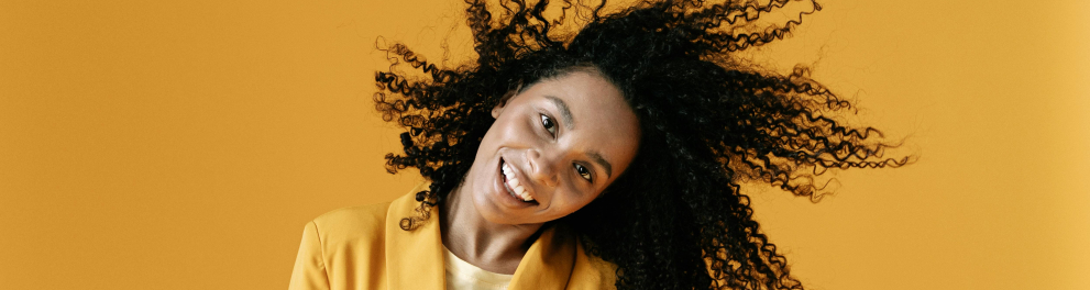 A imagem mostra uma jovem negra em frente a uma parede amarela balançando a cabeça 