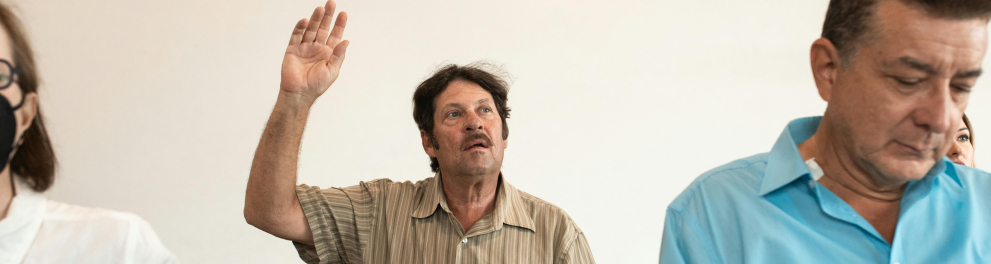 A imagem mostra um idoso em uma sala de aula com uma das mãos levantada