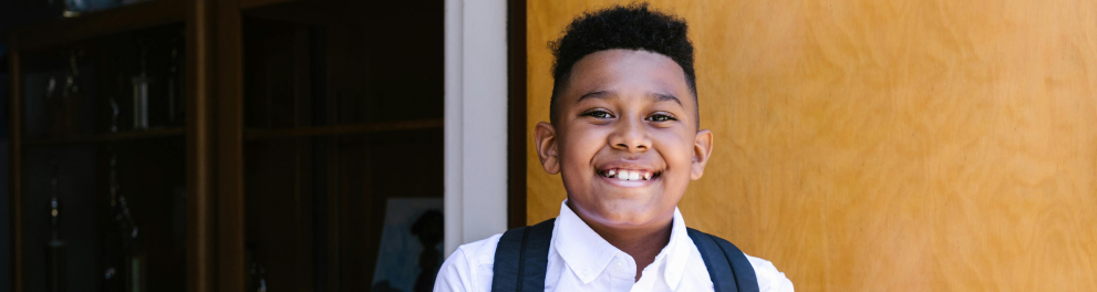 A imagem mostra uma criança negra com uma mochila nas costas e sorrindo para a foto