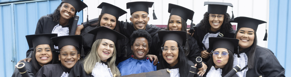 Um grupo diverso de formandos posa sorridente para a foto, usando becas e segurando diplomas. No centro, uma professora também sorri enquanto os abraça. Duas formandas à frente exibem uma placa com a frase: “Melhores professores? Temos.” O clima é de alegria, união e celebração pela conquista.