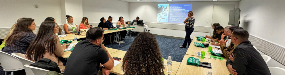 Participantes sentados em mesas organizadas em formato de “U” acompanham uma atividade formativa em uma sala de reunião iluminada. Uma facilitadora fala diante de uma apresentação projetada na parede, enquanto o grupo observa e faz anotações.