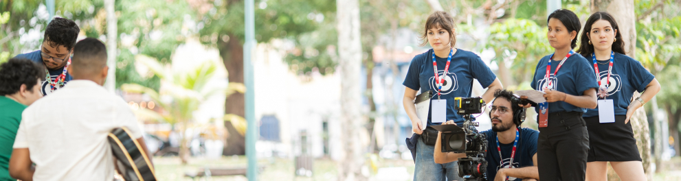 Um grupo de jovens realiza uma gravação ao ar livre, em um parque arborizado. À direita, uma equipe de audiovisual veste camisetas azuis e crachás de identificação; um dos integrantes está agachado atrás de uma câmera profissional montada em tripé, enquanto as colegas acompanham atentamente a cena. À esquerda, em primeiro plano, duas pessoas sentadas em um tronco conversam, uma delas segurando um violão, sugerindo uma entrevista ou apresentação musical sendo registrada.