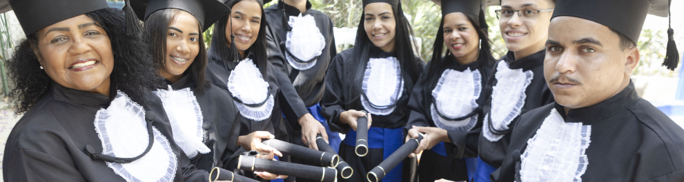 Um grupo de oito formandos posa ao ar livre, todos usando beca preta, faixa azul e capelo (chapéu de formatura). Eles formam um semicírculo, olhando para a câmera e sorrindo, enquanto estendem para frente os canudos de formatura. Ao fundo, há árvores e vegetação, indicando que a foto foi tirada em uma área externa e iluminada.