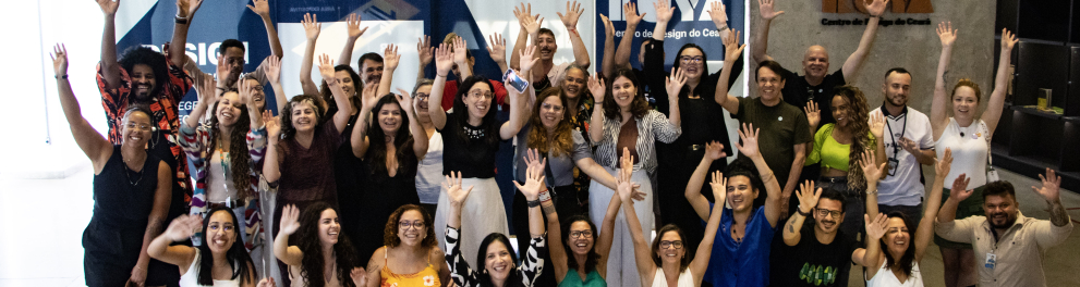 Fotografia em ambiente interno, mostrando um grande grupo de cerca de 40 pessoas reunidas em frente a um painel institucional. Todas estão sorrindo e com os braços levantados para o alto, em gesto de celebração. O grupo é diverso em gênero, idade e tons de pele. As pessoas estão organizadas em várias fileiras: algumas em pé ao fundo, outras agachadas ou sentadas na frente. As roupas são variadas e coloridas, incluindo vestidos, camisetas, calças, sapatênis e sandálias. Ao fundo, há um grande painel com logo