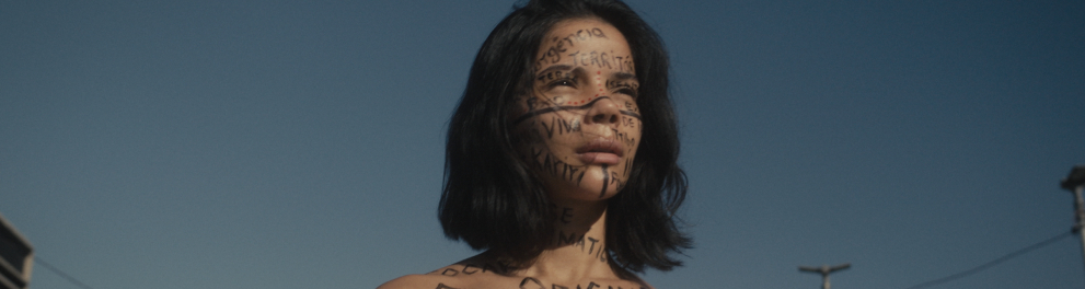 Uma mulher jovem aparece em primeiro plano, ao ar livre, com o céu azul ao fundo. Ela tem cabelo escuro, curto, na altura do queixo, e olha para longe, com expressão séria e reflexiva. Seu rosto, pescoço e parte do tronco estão cobertos por palavras e frases escritas à mão em tinta preta e vermelha, que remetem a temas como violência, invasão, floresta, origem e resistência. Ela veste apenas um top claro, deixando os escritos visíveis no corpo.