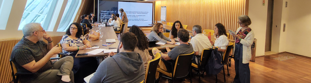 Sala de reunião retangular, com piso de madeira e iluminação amarela no teto. Duas fileiras de mesas formam um grande retângulo, com pessoas sentadas em grupos menores, conversando e escrevendo em cadernos e laptops. Ao fundo, há um telão ligado com um slide que fala em “Diálogo em 3 grupos organizados por eixos” e um quadro branco ao lado. À direita da imagem, uma mulher de cabelos curtos e óculos está em pé, lendo ou anotando em um caderno, enquanto observa os grupos em atividade.
