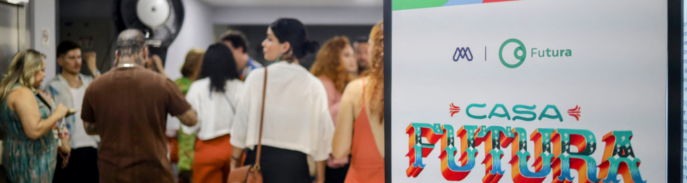 Em primeiro plano e à direita domina um grande cartaz vertical com cores vibrantes e um texto em português. O topo do cartaz tem faixas geométricas coloridas (rosa, amarelo, azul, verde, vermelho) e os logotipos de duas organizações: um símbolo circular roxo ou azul e, ao lado, o logotipo "Futura" em verde. O centro do cartaz tem a frase "CASA FUTURA" em letras grandes, coloridas e estilizadas, e abaixo, em letras menores, o slogan: "O FUTURO COMEÇA AGORA". A parte inferior do cartaz repete o padrão geométr