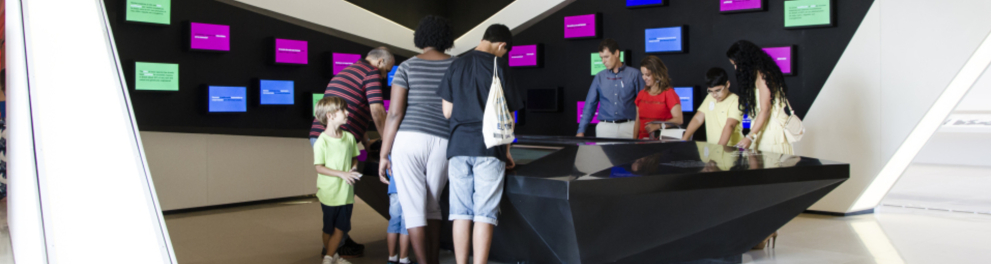 Imagem de uma sala de museu com uma mesa, adultos e crianças ao redor