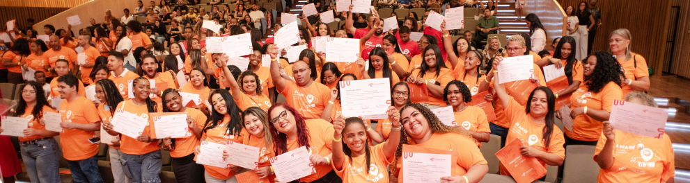 Participantes de uma cerimônia de certificação posam reunidos no auditório, segurando seus certificados com entusiasmo. Os formandos, vestidos com camisetas laranja, ocupam a frente da sala, enquanto o público acompanha a celebração nas arquibancadas ao fundo. A cena transmite sentimento de orgulho coletivo e comemoração.