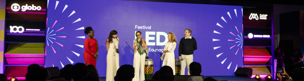 Em um palco de evento, cinco pessoas estão em pé, alinhadas, diante de uma plateia numerosa sentada. O público aparece em primeiro plano, visto de costas e em silhueta. No centro do palco, uma mulher fala ao microfone; à sua frente há dois tambores altos, decorados com padrões coloridos. O cenário ao fundo é um grande painel roxo com a inscrição “Festival LED Educação”, acompanhado de elementos gráficos que lembram raios ou explosões coloridas nas laterais.