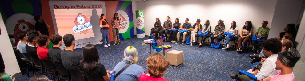 Sala ampla com cadeiras organizadas em círculo, onde cerca de 30 jovens participam de um encontro do Geração Futura 2026. No centro, duas mediadoras conduzem a atividade, enquanto os participantes escutam atentamente. Ao fundo, um painel colorido com a identidade visual do projeto e do Canal Futura. O ambiente transmite troca, escuta e formação coletiva em produção audiovisual.