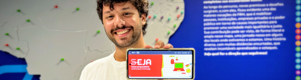 Um homem jovem sorri para a câmera enquanto segura um celular com a tela voltada para a frente. Ele usa camiseta branca e está em pé diante de um painel azul. No fundo, há um mapa do Brasil em relevo, com vários pequenos pontos coloridos espalhados pelo território. Na tela do celular aparece uma página com fundo vermelho e ilustrações coloridas, com o texto em destaque: “SEJA – Curso preparatório online para o Encceja”