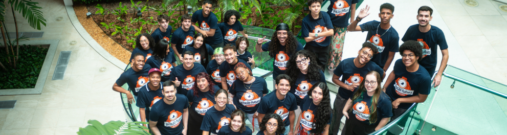 Foto feita de um ponto mais alto, mostrando um grupo de jovens reunidos em uma varanda interna com guarda-corpo de vidro curvo. Todos usam camisetas azul-marinho com o logotipo do projeto “Geração Futura” em destaque no peito e crachás pendurados no pescoço.  O grupo está reunido de forma descontraída, olhando para cima em direção à câmera.