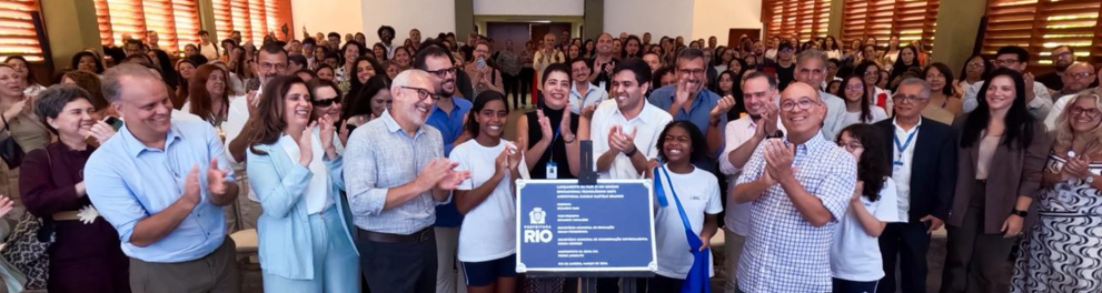 A foto registra um momento de celebração coletiva em um auditório cheio. Autoridades, educadores e estudantes se reúnem ao redor de uma placa comemorativa da Prefeitura do Rio e aplaudem, enquanto muitas pessoas acompanham o momento ao fundo.