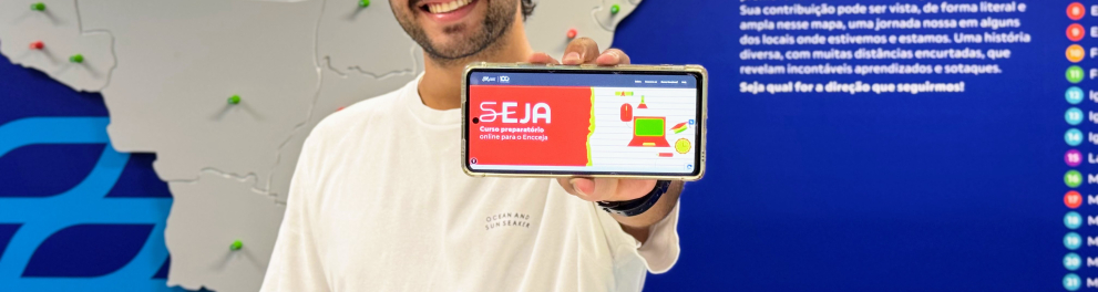 Um homem jovem, sorridente, segura um celular em direção à câmera exibindo a tela do programa SEJA – curso preparatório online para o Encceja. Ele veste camiseta branca e está em frente a um grande mapa do Brasil fixado na parede, marcado com vários pinos coloridos que indicam diferentes localidades.