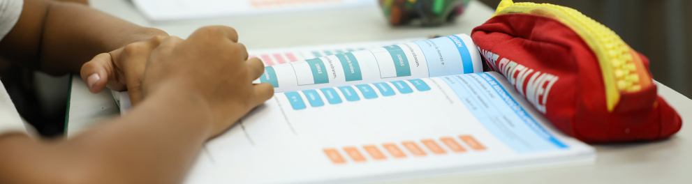 Em sala de aula, um estudante mantém as mãos apoiadas sobre um material didático aberto na mesa. Ao lado, um estojo vermelho e um recipiente com lápis de cor completam o cenário. Ao fundo, outros alunos aparecem desfocados, indicando um ambiente coletivo de aprendizagem. A imagem destaca a rotina escolar, com foco no estudo, na organização dos materiais e no processo de ensino-aprendizagem.