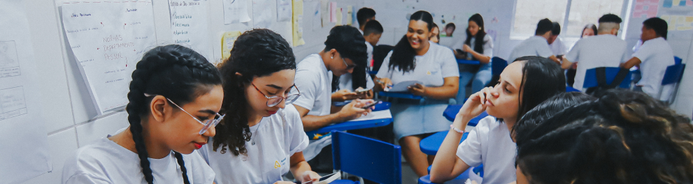 Sala de aula com estudantes sentados em carteiras azuis, organizados em pequenos grupos. A maioria usa camiseta branca e está com celulares nas mãos, aparentemente realizando uma atividade juntos. Há folhas impressas sobre as mesas e cartazes nas paredes ao fundo, indicando um ambiente escolar ativo e colaborativo.