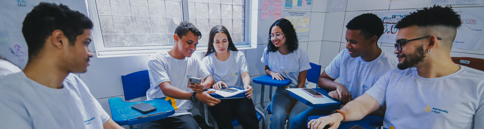 Um grupo de seis jovens está sentado em círculo dentro de uma sala de aula, em cadeiras com apoio para escrita. Todos usam camisetas brancas com um pequeno logotipo colorido. No centro, dois deles compartilham um celular, mostrando algo na tela, enquanto os demais observam com atenção e sorriem. Alguns têm cadernos ou celulares apoiados nas mesas acopladas às cadeiras.